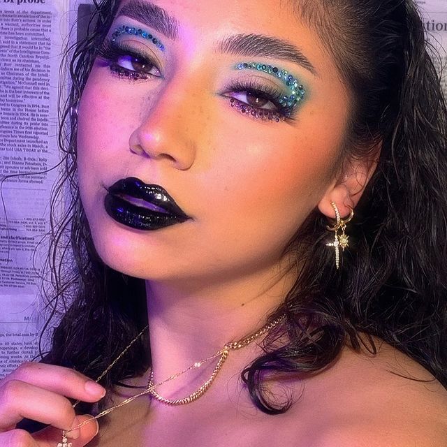 maquiagem-com-batom-preto Selfie de uma mulher. Ela usa o cabelo solto, maquiagem com pedrinhas de glitter no olho, batom preto e colares dourados. Ela olha para a câmera e não sorri.