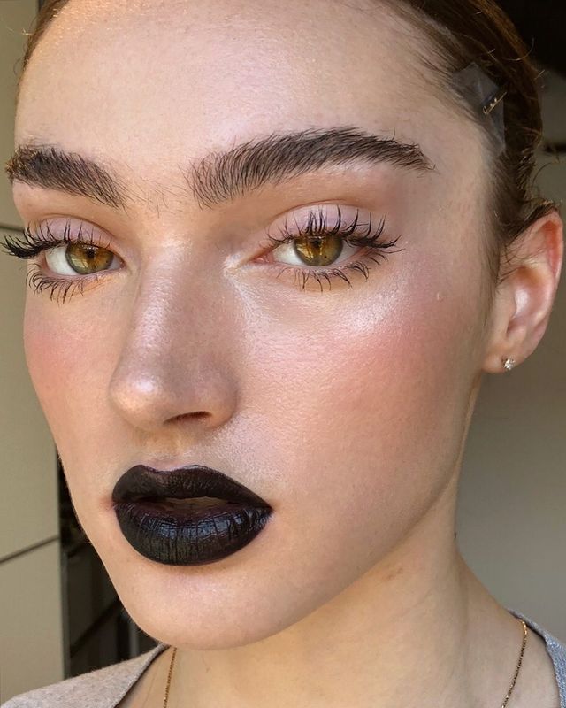 maquiagem-natural-com-batom-preto Foto do rosto de uma mulher. Ela está com o cabelo preso, pele natural, sobrancelhas penteadas, rímel e batom preto. Ela olha para a câmera e não sorri.
