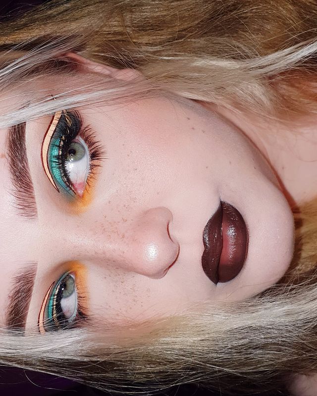 maquiagem-sombra-colorida-batom-escuro Foto do rosto de uma mulher. Ela usa o cabelo solto, delineado colorido nos tons de amarelo e verde, pele natural e batom escuro. Ela olha para o lado e não sorri.