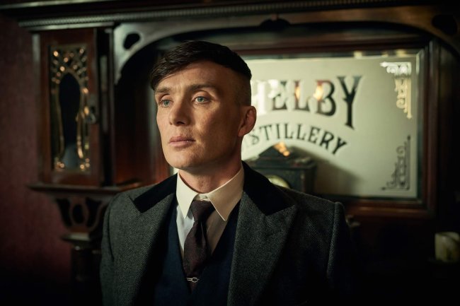 peaky Um homem com terno e sobretudo em frente à um móvel de madeira com um escrito de vidro.