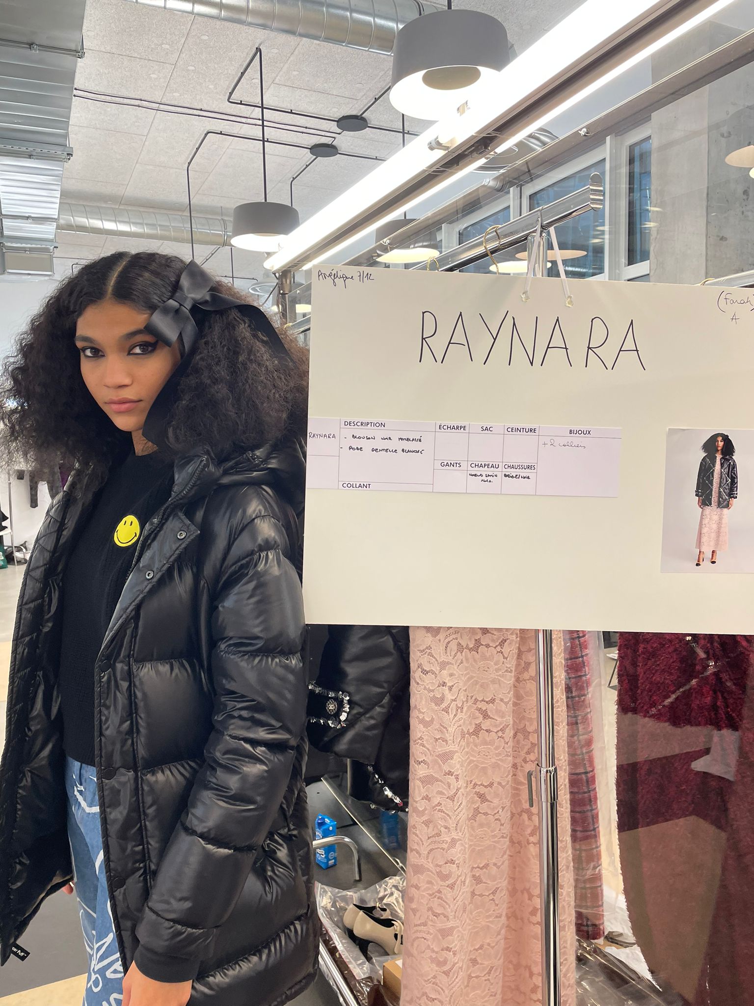 Raynara Negrine para Chanel – Métiers d’art 2021_1 Raynara Negrine nos bastidores do desfile Metiers D'Art da Chanel. Ela posa à esquerda, ao lado de um painel com seu nome e o look que usa no desfile, de jaqueta puffer preta, camiseta preta, calça jeans e laço no cabelo.