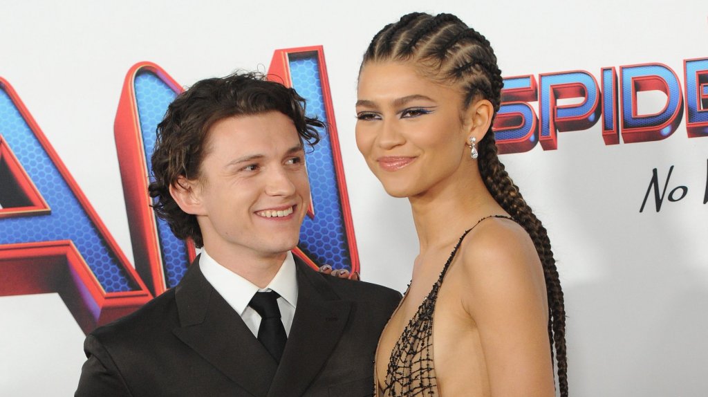 Tom e Zendaya posando para fotos. Ela está com um vestido nude com detalhes em preto e ele está de terno preto e camisa branca.