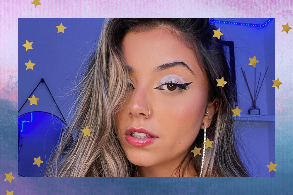 Selfie de Isa Paoli olhando para câmera com expressão séria; ela usa uma maquiagem com delineado preto, sombra roxa e gloss rosa; a parede ao fundo é azul; a margem é uma textura de degradê em tons de roxo, rosa, azul e branco com estrelas amarelas como decoração