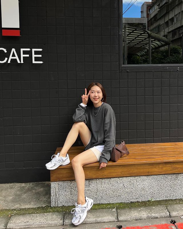 look-casual-com-tenis-esportivo Foto de uma mulher sentada em um banco em frente a uma cafeteria. Ela usa um moletom cinza oversized, short branco, bolsa baguete marrom e tênis esportivo branco. Ela apoia a perna direita no banco, faz sinal de