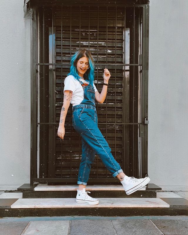 Foto de uma mulher em frente a uma porta. Ela usa uma camiseta branca, macacão jeans e tênis branco de cano alto. Ela olha para baixo e sorri.