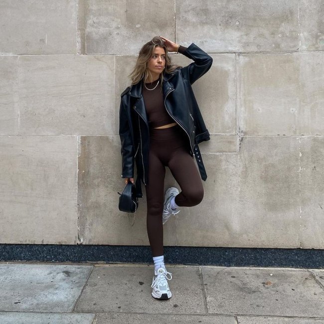 look com tenis esportivo Foto de uma mulher em uma calçada. Ela usa um top cropped marrom, calça legging marrom, jaqueta de couro preta, bolsa de couro preta e tênis branco esportivo com meia longa. Ela está com o pé esquerdo apoiado na parede, mão esquerda no cabelo, olha para o lado e não sorri para a foto.