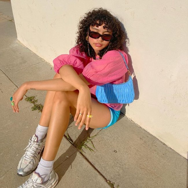 look-com-tenis-esportivo Foto de uma mulher sentada no chão. Ela usa uma blusa rosa de manga bufante, short azul, bolsa baguete azul, óculos de sol e tênis esportivo branco. Ela olha para o lado e não sorri para foto.