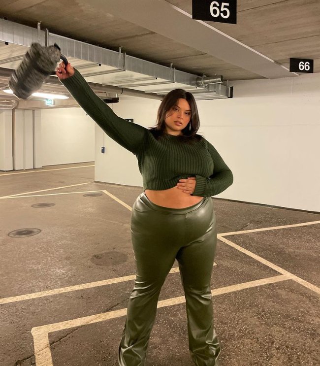 look-verde-monocromatico Foto de uma mulher em um estacionamento. Ela usa um top cropped de manga longa verde, calça de couro verde no mesmo tom e uma bolsa cinza. Ela está com a mão esquerda na barriga, mão direita levantada, olha para a câmera e não sorri.