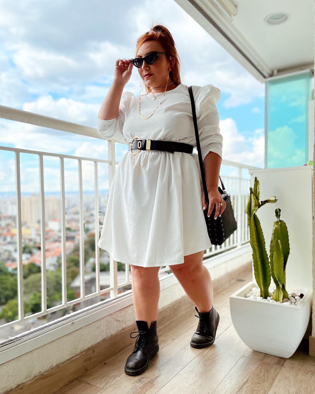 Garota usando vestido branco com cinto preto, botinha preta, bolsa e óculos de sol pretos. Seu cabelo está preso em um rabo e ela está na varanda de um apartamento