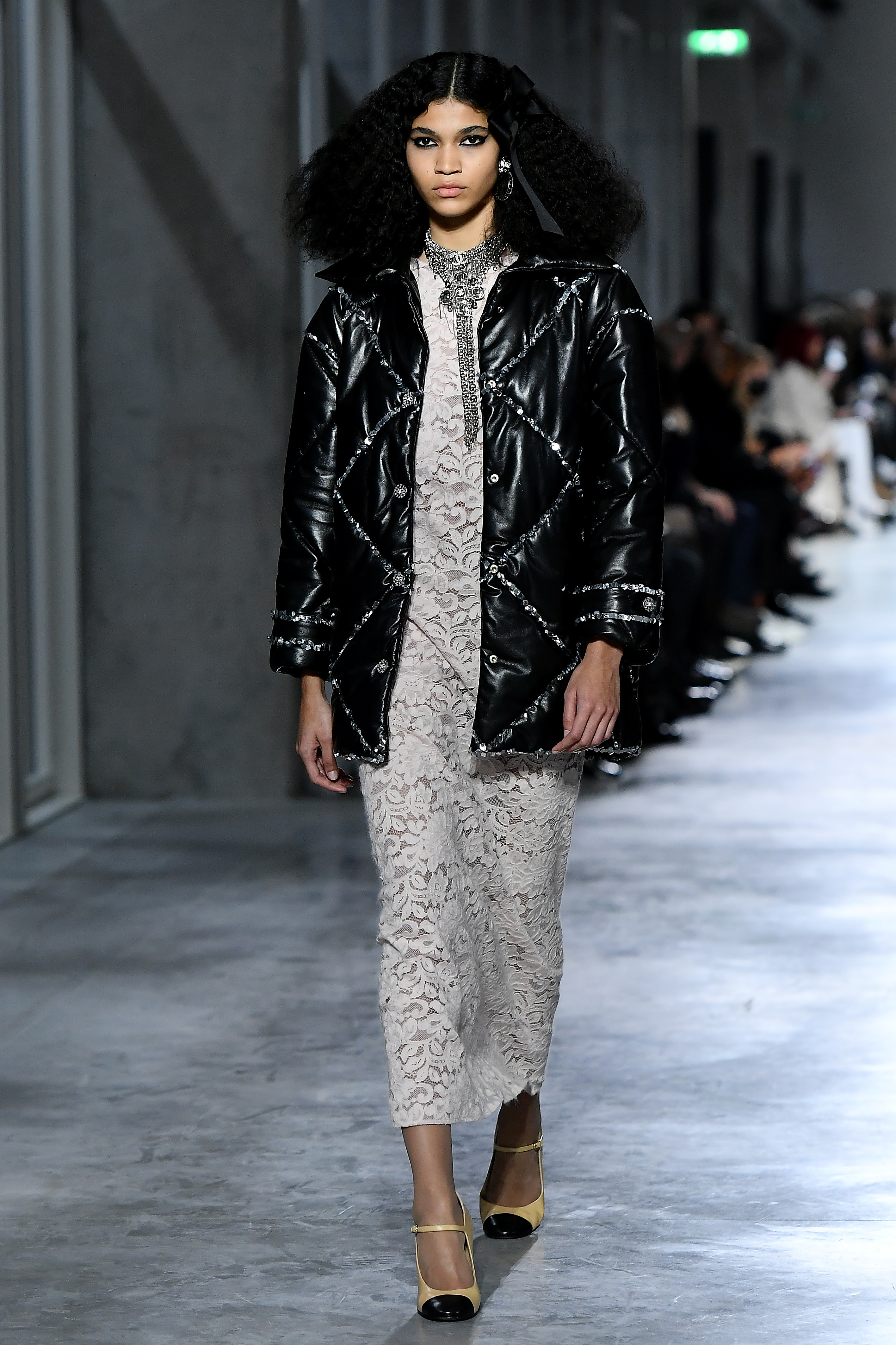 Chanel Metiers D’Art 2021-2022 : Runway At Le 19M Raynara Negrine usando vestido longo de renda com jaqueta puffer preta por cima no desfile Metiers D'Art 2021-2022 da Chanel