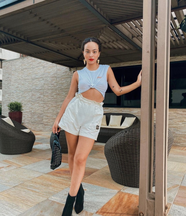 regata-com-no Foto de uma mulher em uma casa. Ela usa uma regata branca com nó na frente, short branco, bota preta e pochete preta. Ela está com o cabelo preso, maquiagem com batom vermelho, olha para a câmera e não sorri.