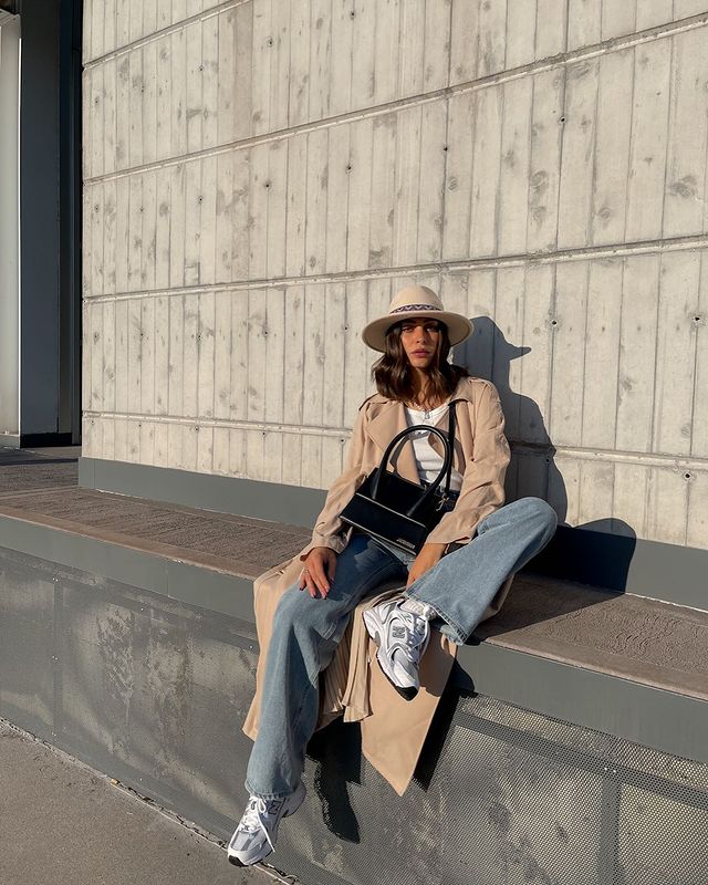 tenis-casual-look Foto de uma mulher sentada em uma calçada alta. Ela usa uma camiseta branca, sobretudo longo bege, calça jeans, tênis branco esportivo, bolsa preta e chapéu bege. Ela olha para a câmera e não sorri.