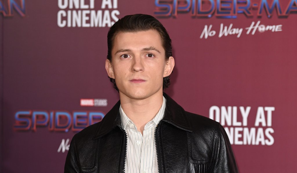 Tom Holland posando em tapete vermelho, com jaqueta de couro preta, calça preta e camisa branca.