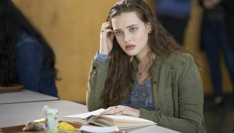 Foto de Hannah Baker, de 13 Reasons Why, sentada numa carteira de sala de aula, com um livro aberto sobre a mesa, com a mão direita no cabelo e olhando para frente com ar curioso