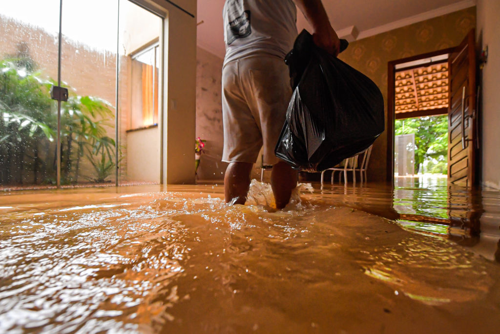 Foto de um senhor, de costas, carregando um saco de lixo. Ele está na sala de casa e a água da enchente chega até os tornozelos