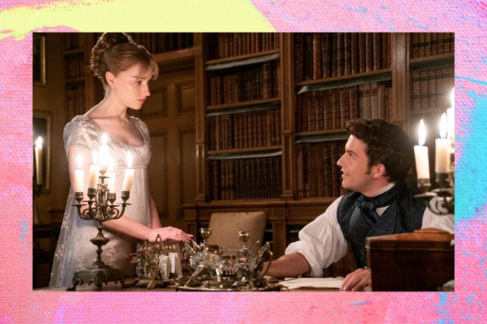 Foto dos bastidores de Bridgerton. Uma mulher de coque e vestido ao lado de uma mesa com um homem sentado.