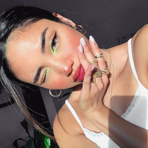 maquiagem-canto-interno-neon-rachelteetyler Selfie de uma mulher. Ela usa uma blusa de alcinha branca, cabelo preso, brinco de argola e maquiagem simples com detalhe neon no canto interno dos olhos.