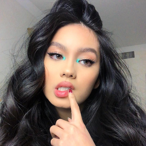 neon-azul-canto-interno-olho-jessyluxe Selfie de uma mulher. Ela está com o cabelo solto e uma maquiagem simples com delineador gatinho e ponto de luz azul neon no canto interno dos olhos. Ela está com o dedo indicador esquerdo na boca, olha para o lado e não sorri para a foto.