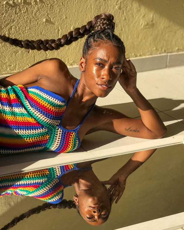 cabelo-trancado-com-rabo-de-cavalo Foto de uma mulher deitada com a cabeça apoiada na mão esquerda. Ela usa uma blusa de alcinha de crochê e está com o cabelo preso em um rabo de cavalo alto e trançado. Ela olha para a câmera e não sorri.