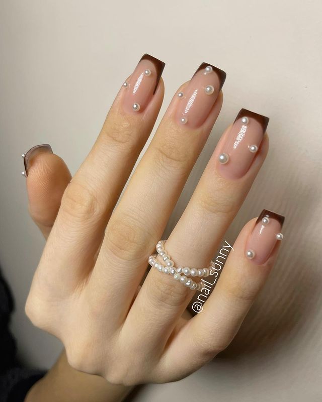 francesinha-marrom-com-perolas Foto de uma mão com as unhas decoradas com francesinha marrom e aplicação de pérolas.