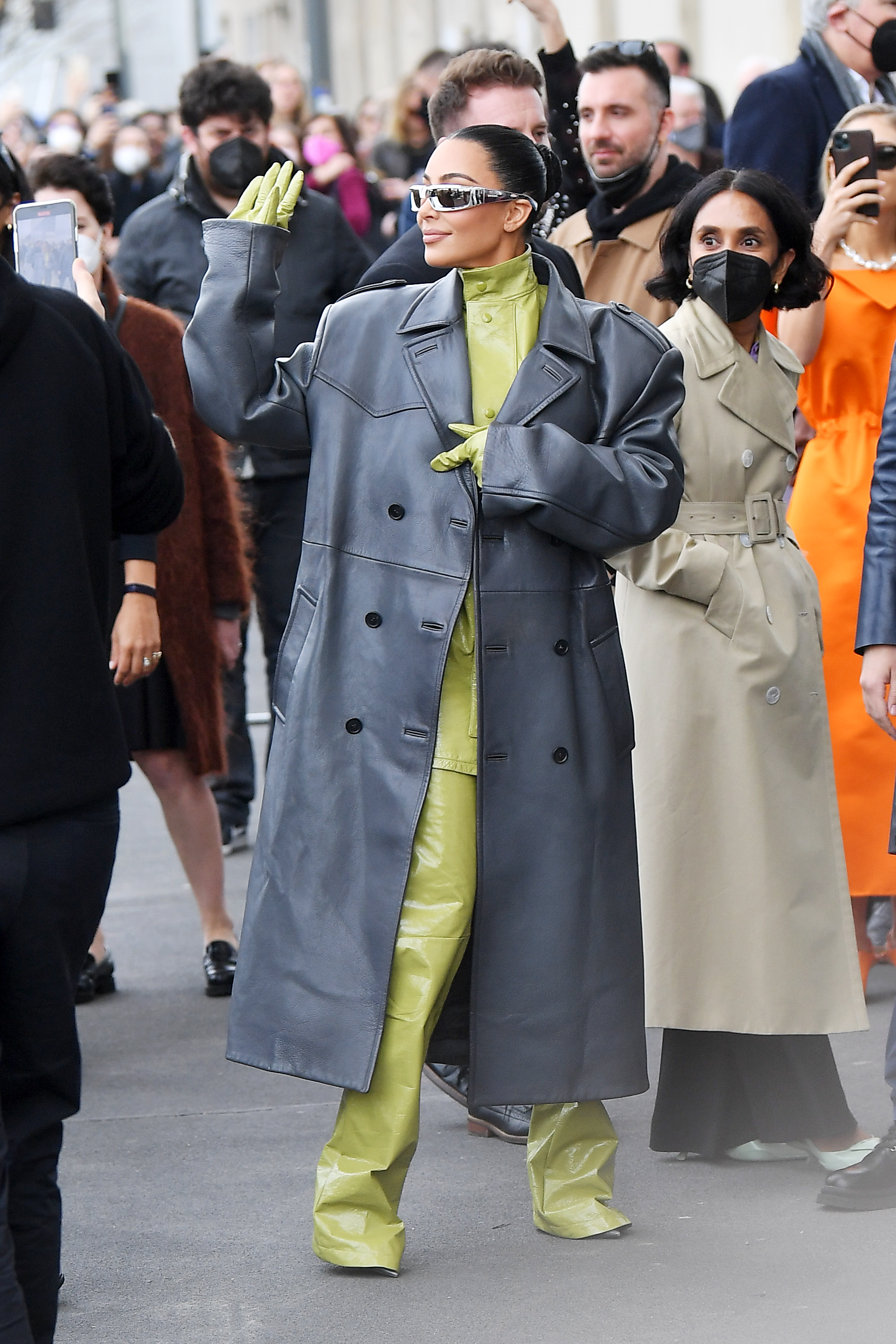 Prada – Outside Arrivals – Milan Fashion Week Fall/Winter 2022/2023 Kim Kardashian na semana de moda de Milão usando macacão verde com sobretudo cinza e óculos escuros. Ela olha para o lado e levanta a mão para cumprimentar fãs