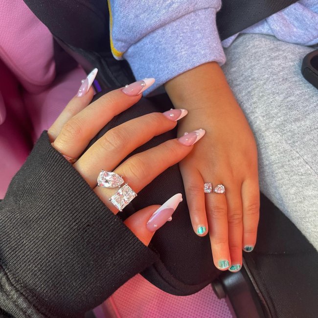kylie-jenner-unhas-francesinha-com-perolas Foto das mãos da Kylie Jenner e sua filha Stormi. A Kylie está com as unhas longas com uma decoração de francesinha branca e pérolas e ambas usam anéis combinando.