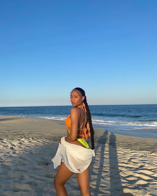 tranca-no-rabo-de-cavalo Foto de uma mulher na praia. Ela usa um biquíni laranja, segura uma camisa branca e está com um penteado de rabo de cavalo baixo trançado. Ela está de lado, olha para a câmera e não sorri para a foto.