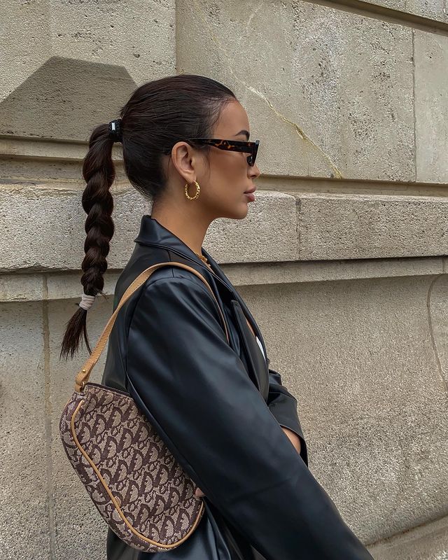 Foto de uma mulher de perfil em uma calçada. Ela usa uma jaqueta de couro preta, bolsa baguete marrom, óculos de sol preto, brinco de argola dourado e penteado rabo de cavalo alto trançado. Ela olha para frente e não sorri para a foto.