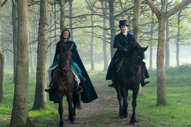 BRIDGERTON_201_Unit_06300R Simone Ashley como Kate Sharma e Jonathan Bailey como Anthony Bridgerton em cena de Bridgerton; eles estão andando de cavalo lado a lado em uma trilha na floresta enquanto sorriem