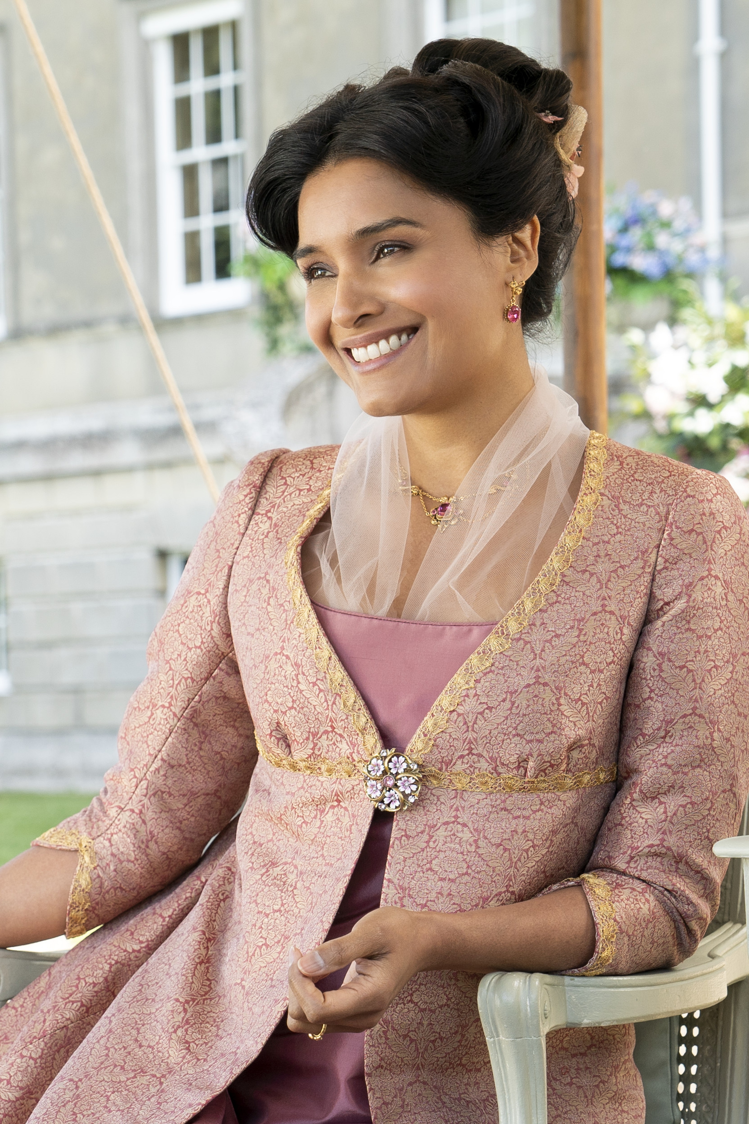 Imagem mostra Shelley Conn caracterizada como Mary Sharma, sentada e sorrindo em cena da 2ª temporada de Bridgerton.