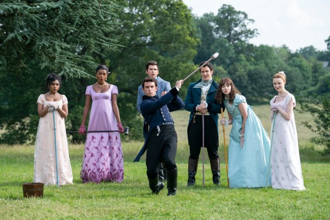 BRIDGERTON_203_Unit_04220R Charithra Chandran como Edwina Sharma, Simone Ashley como Kate Sharma, Luke Newton como Colin Bridgerton, Luke Thompson como Benedict Bridgerton, Jonathan Bailey como Anthony Bridgerton, Claudia Jessie como Eloise Bridgerton, Phoebe Dyvenor como Daphne Basset em cena na segunda temporada de Bridgerton; eles estão em uma área verde aberta observando Anthony fazer uma jogada com um taco de Pall Mall