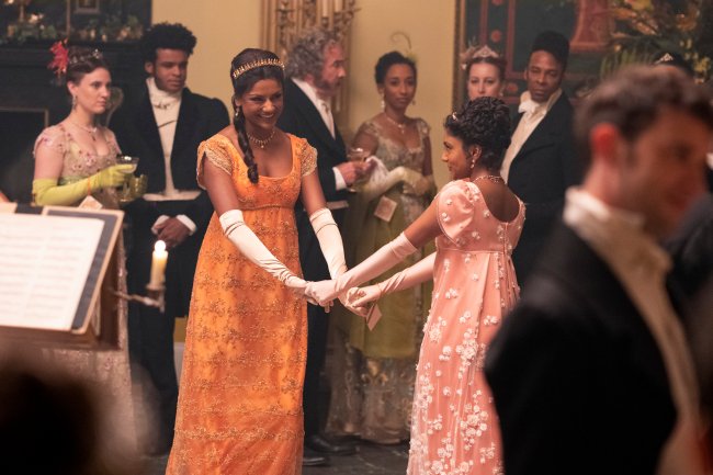 BRIDGERTON_208_Unit_00518R Simone Ashley como Kate Sharma e Charithra Chandran como Edwina Sharma em cena de Bridgerton; as duas estão da mãos dadas dançando em um baile no salão enquanto dão risada