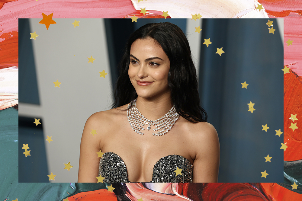 Imagem mostra Camila Mendes no tapete azul da Vanity Fair Oscar Party 2022 usando um vestido tomara que caia prateado e uma maquiagem clássica. O registro segue acompanhado de uma borda com pinceladas de tinta e estrelinhas douradas por toda imagem.