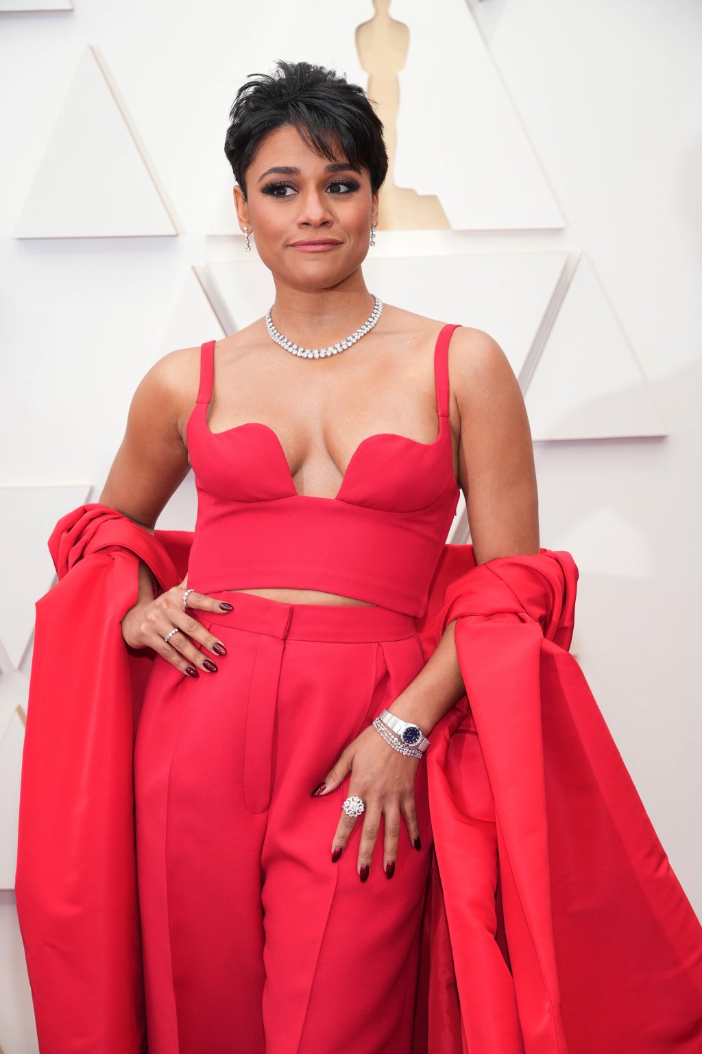 Ariana DeBose no Oscar 2022; ela está posando com a mão na cintura com um look vermelho com colar de brihantes enquanto sorri levemente