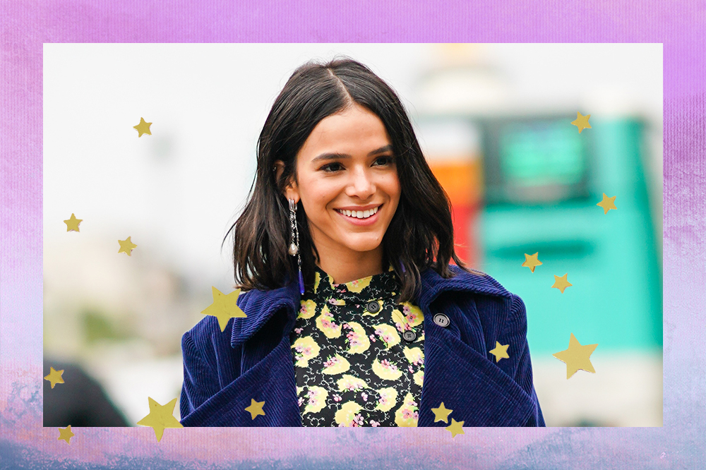 Montagem de foto de Bruna Marquezine em fundo degradê lilás. Ela está sorrindo e olhando para o lado, usando casaco azul de veludo e vestido estampado