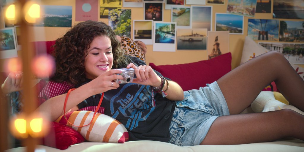 Anita (Maisa Silva) na série De Volta aos 15 deitada em sofá com camiseta de banda, short jeans e meia-calça com uma câmera digital nas mãos