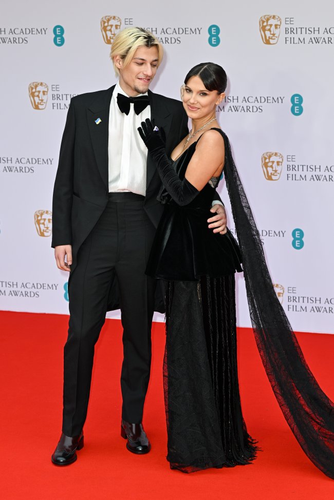 EE British Academy Film Awards 2022 – Red Carpet Arrivals Jake Bongiovi e Millie Bobby Brown no tapete vermelho do British Academy Film Awards 2022, em Londres posando para foto e sorrindo levemente; ele está abraçando a atriz pela cintura enquanto a olha