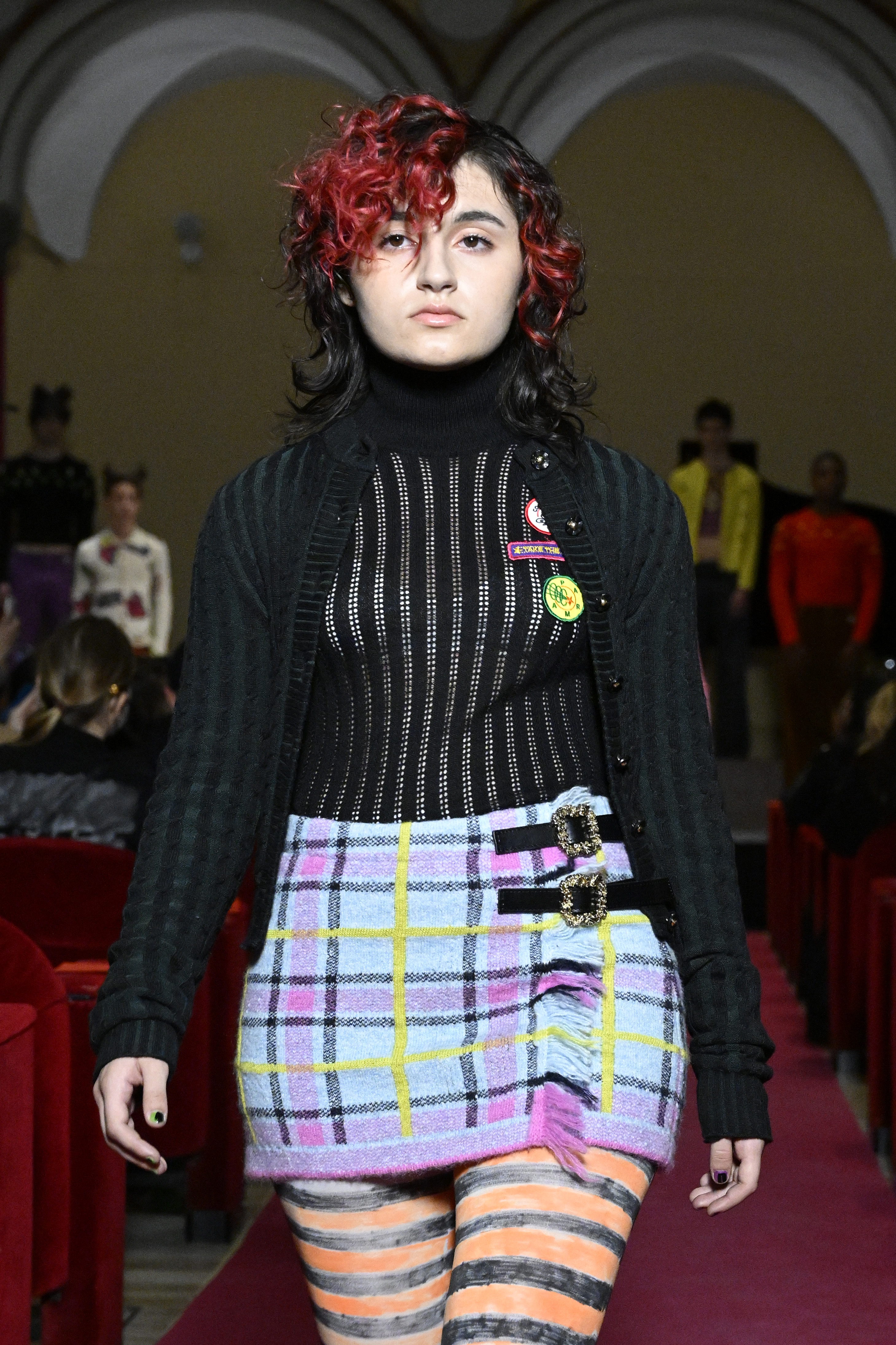 Cormio – Runway – Milan Fashion Week Fall/Winter 2022/2023 Modelo com cabelo preto e vermelho, tricô de manga comprida preto e saia xadrez em desfile da Cormio na semana de moda de Milão