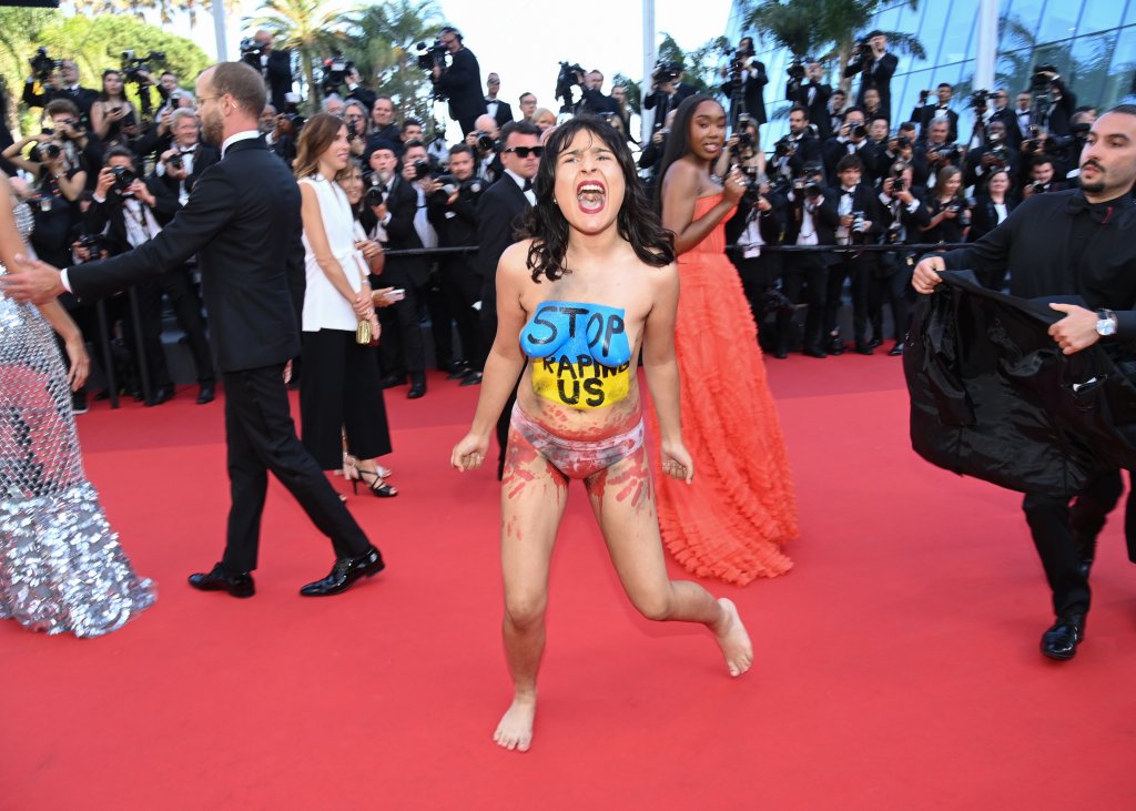 Foto de uma mulher seminua, apenas de calcinha, protestando no tapete vermelho do Festival de Cannes. Ela está com a bandeira da Ucrânia pintada no peito, sob os dizeres "Pare de nos estuprar"