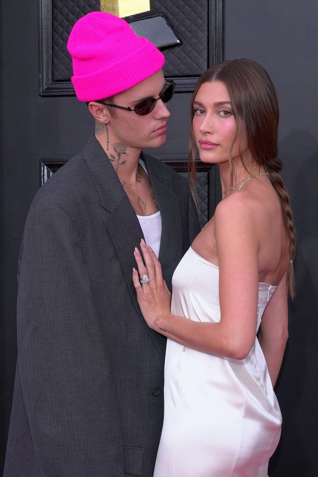 hailey-bieber-trancas-boho-grammy-2022 Foto Justin e Hailey Bieber no tapete vermelho do Grammy 2022. Na imagem Justin usa uma touca rosa, blazer oversized cinza, regata branca e óculos escuros. Hailey usa um vestido branco tomara que caia, maquiagem natural e cabelo preso em uma trança boho despojada.