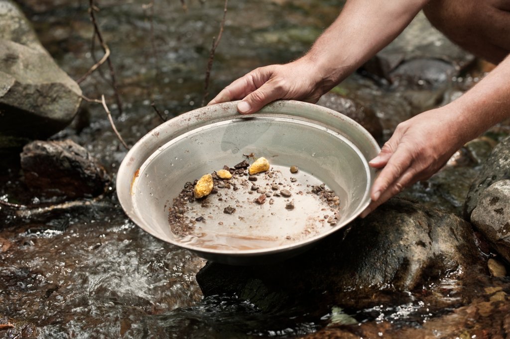 Duas mãos segurando uma tigela de prata com pedaços de ouro e terra dentro