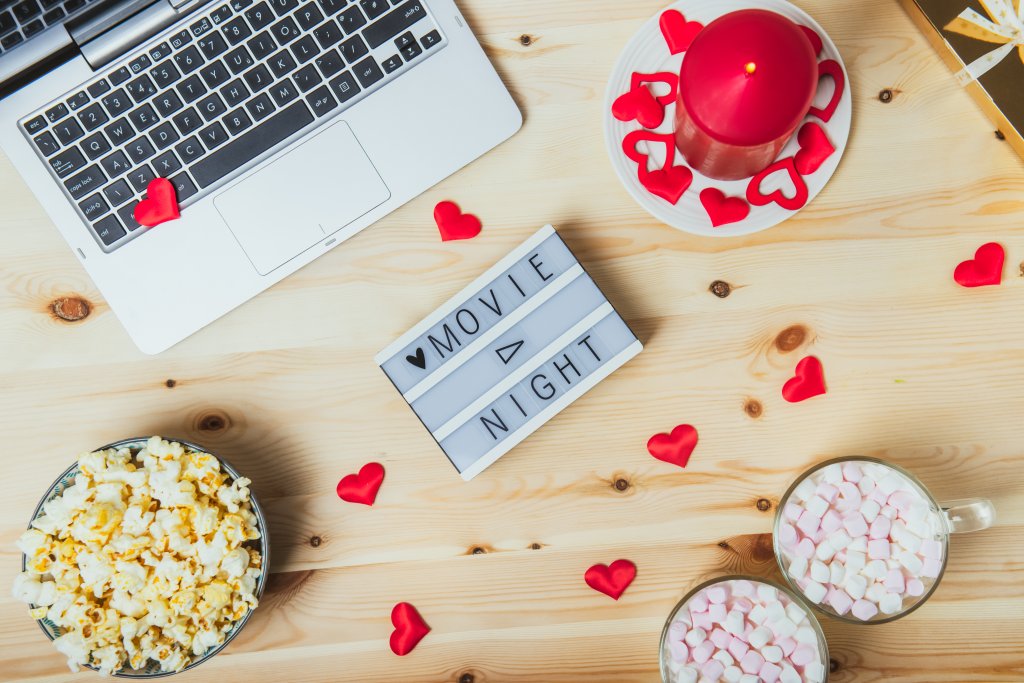 Foto de uma mesa em tom de madeira clara, com um notebook, pipoca, corações vermelhos espalhados e letreiro escrito "movie