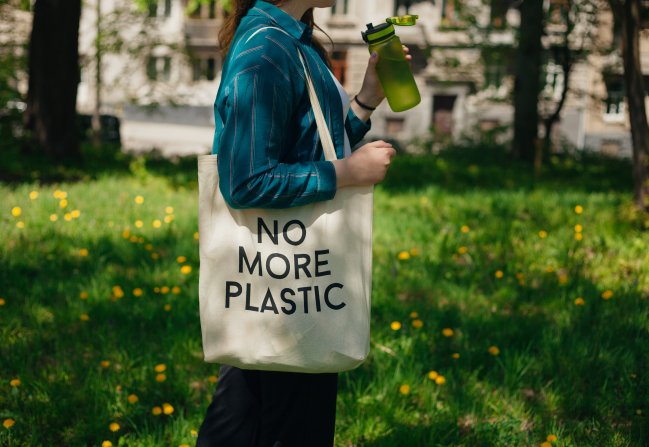 Uma mulher bebendo água direto de uma garrafa reutilizável e segurando uma ecobag que diz 