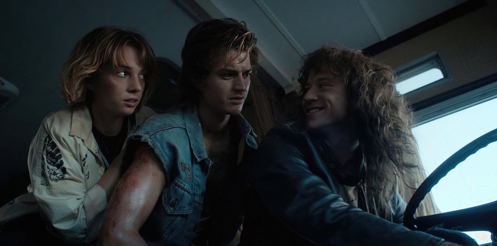 Maya Hawke como Robin Buckley, Joe Keery como Steve Harrington e Joseph Quinn como Eddie Munson em Stranger Things; eles estão em um trailer e Eddie está olhando para Steve sorrindo enquanto Steve e Robin olham para ele com expressões confusas
