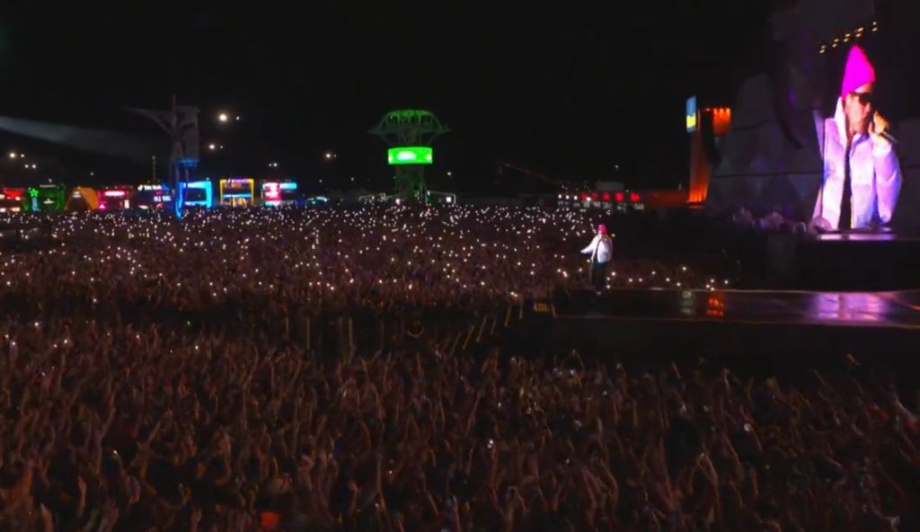 Público assistindo show de Justin Bieber; ele aparece no palco cantando com casaco roxo e touca rosa; ao fundo, é possível ver o cantor no telão usando óculos escuros enquanto segura o microfone e olha para os fãs