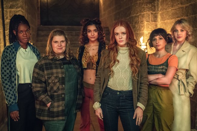 Precious Mustapha como Aisha, Eliot Salt como Terra, Paulina Chavez como Flora, Abigail Cowen como Bloom, Elisha Applebaum como Musa e Hannah Van Der Westhuysen como Stella posando para foto com expressões sérias e neutras com uma parede de tijolos e luzes com fogo em tochas