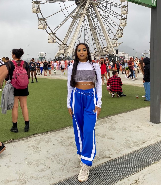 Foto de uma menina no Rock in Rio 2022. Ela usa uma blusa de manga longa branca, top de brilhos como sobreposição e calça azul. Ela olha para a câmera e não sorri.