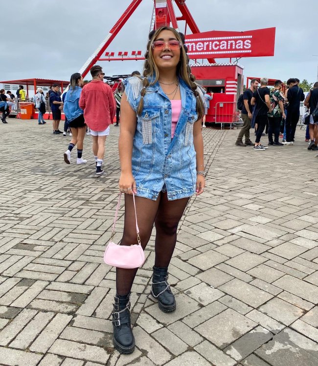 Foto de uma menina no Rock in Rio. Ela usa uma salopete jeans, meia calça e coturno. Ela olha para a câmera e sorri.