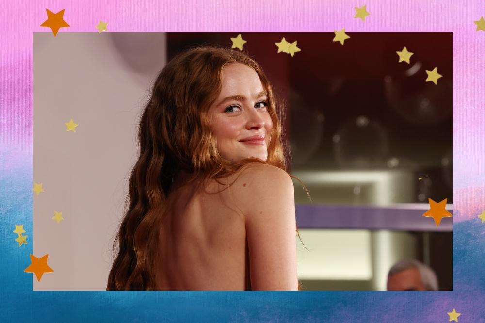 Montagem com o fundo colorido em um degradê de roxo e azul com detalhe de estrelas coloridas nas bordas com a foto da atriz Sadie Sink no centro. Na foto ela está de perfil e cabelo solto, olha para a câmera e sorri levemente.