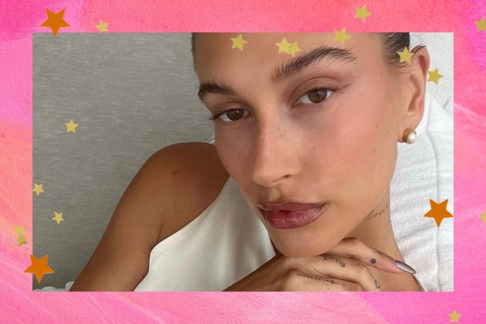 Montagem com o fundo rosa com estrelas douradas nas bordas com uma selfie do rosto da Hailey Bieber no centro. Ela olha para a câmera e não sorri.